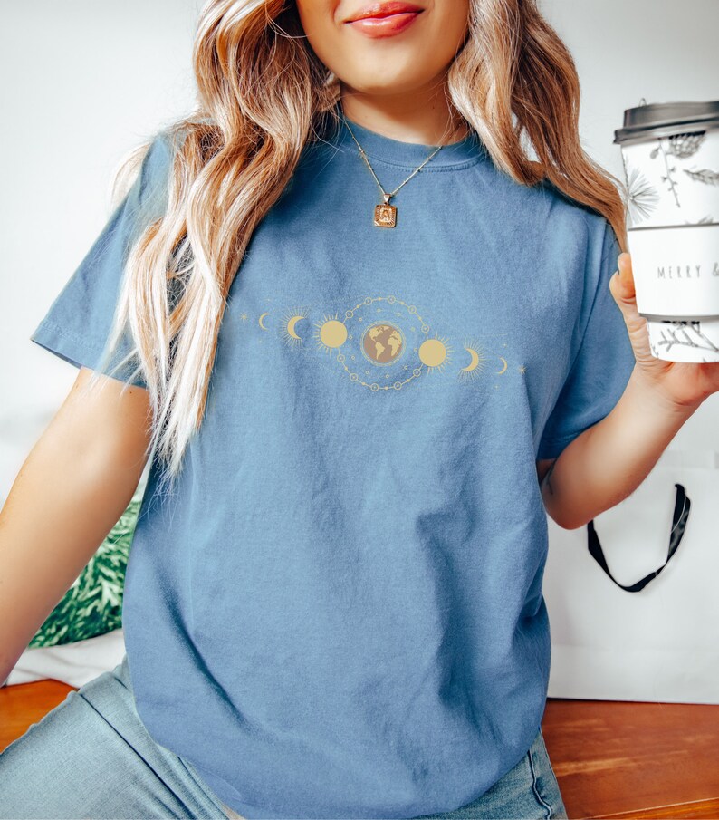 K&ouml;nnte beinhalten: Ein blaues T-Shirt mit einem Aufdruck einer stilisierten Erde, umgeben von Sonnen und Monden. Der Aufdruck ist in Gold und Gelb.