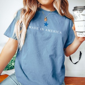 K&ouml;nnte beinhalten: Ein blaues T-Shirt mit einem wei&szlig;en Stern und dem Text "MADE IN AMERICA" auf der Vorderseite.
