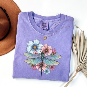 May include: A lavender Comfort Colors t-shirt featuring a dragonfly design with blue, pink, and yellow flowers. The dragonfly has blue and green wings. A gold necklace is draped on the shirt.