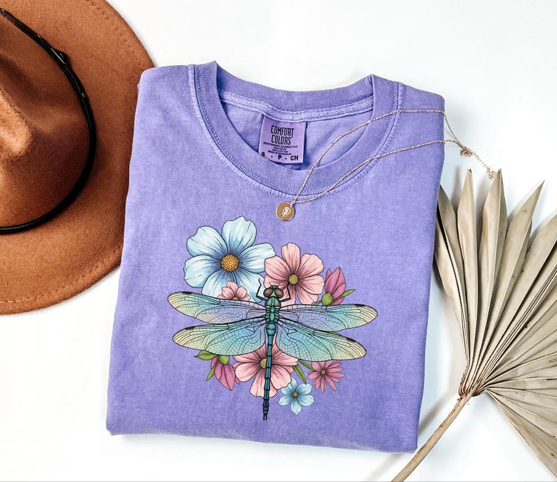 May include: A lavender Comfort Colors t-shirt featuring a dragonfly design with blue, pink, and yellow flowers. The dragonfly has blue and green wings. A gold necklace is draped on the shirt.