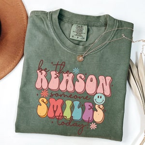 May include: Green t-shirt with a retro-style graphic that says "Be the reason someone smiles today!" in colorful letters with a smiley face.