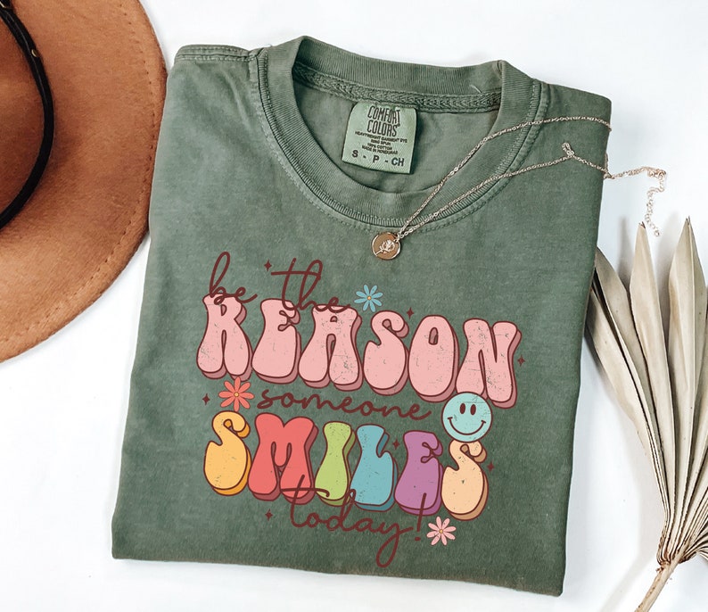 May include: Green t-shirt with a retro-style graphic that says "Be the reason someone smiles today!" in colorful letters with a smiley face.