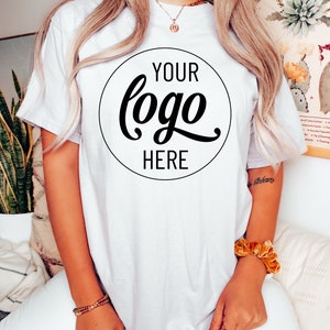 May include: White t-shirt with a black circle on the front that says "YOUR logo HERE" in a cursive font.