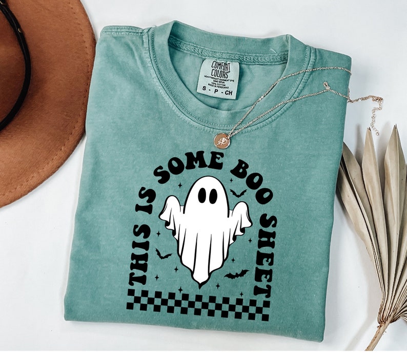 以下が含まれることがあります： 白の幽霊のグラフィックと「This is some boo sheet」というテキストが書かれたターコイズブルーのTシャツです。
