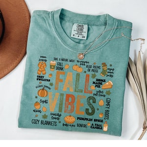 May include: A teal blue t-shirt with a graphic design featuring the text "Fall Vibes" in a vintage style. The design includes various fall-themed illustrations such as pumpkins, leaves, cozy sweaters, and a pie. The text "Comfort Colors" is visible on a tag attached to the shirt.