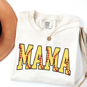 May include: A white t-shirt with the word "MAMA" printed in yellow with red stitching, resembling a softball. The shirt has a tag that reads "Comfort Colors".