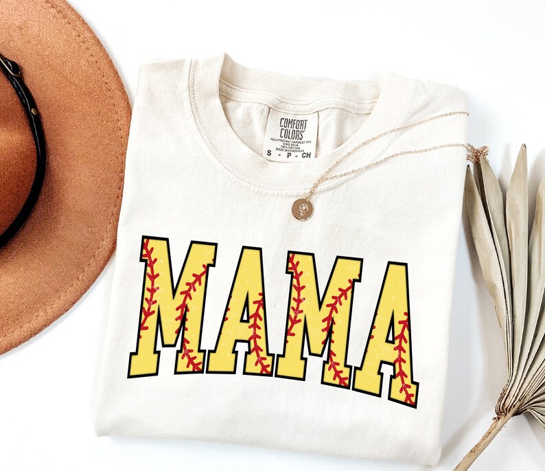 May include: A white t-shirt with the word "MAMA" printed in yellow with red stitching, resembling a softball. The shirt has a tag that reads "Comfort Colors".