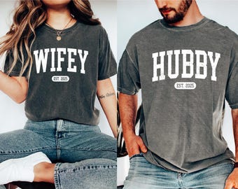 Puede incluir: Dos camisetas grises con texto blanco. La primera camiseta dice "WIFEY EST. 2025" y la segunda camiseta dice "HUBBY EST. 2025".