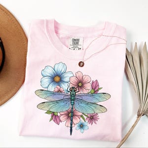 May include: A light pink t-shirt featuring a colorful dragonfly design with blue, pink, and green hues, surrounded by flowers. The shirt has a crew neck and short sleeves. A gold necklace with a pendant is on the shirt.