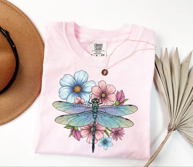 May include: A light pink t-shirt featuring a colorful dragonfly design with blue, pink, and green hues, surrounded by flowers. The shirt has a crew neck and short sleeves. A gold necklace with a pendant is on the shirt.