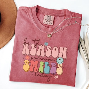 May include: A red t-shirt with the text "be the reason someone smiles today!" in a retro, colorful font. The text is surrounded by small flowers and a smiley face.