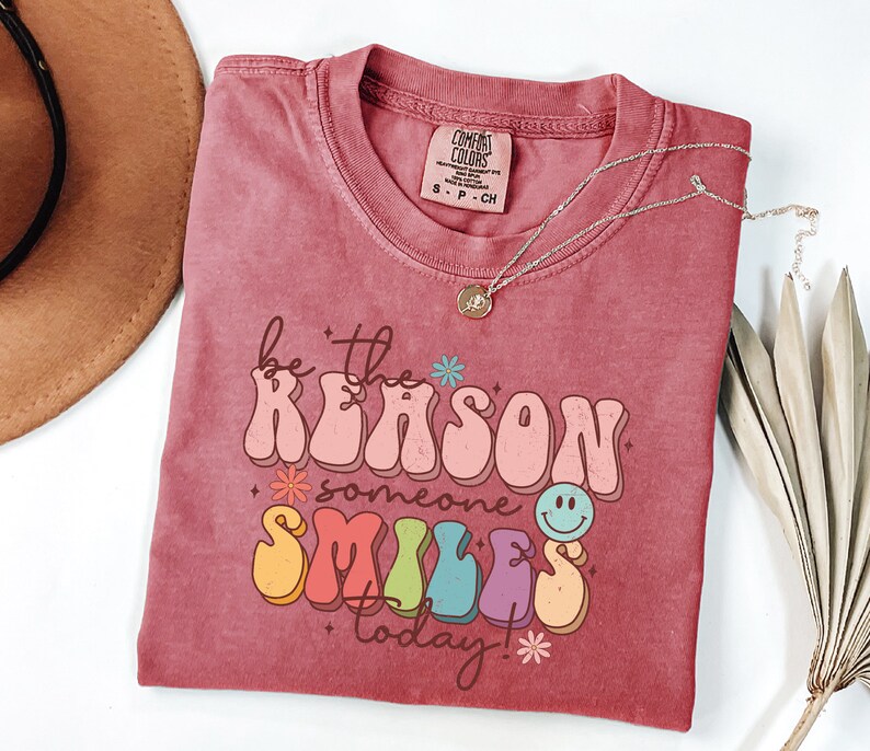 May include: A red t-shirt with the text "be the reason someone smiles today!" in a retro, colorful font. The text is surrounded by small flowers and a smiley face.