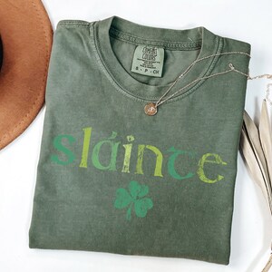May include: A green t-shirt with the word "sl&aacute;inte" in a gradient of green and yellow. A four-leaf clover is below the word.