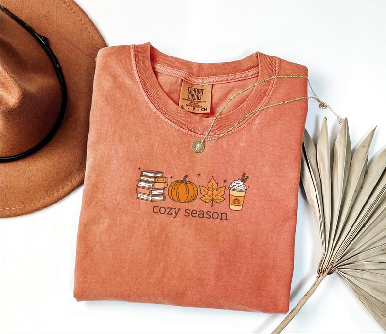 May include: A coral-coloured t-shirt with a crew neck and the words "cozy season" printed on the front. The graphic includes a stack of books, a pumpkin, a maple leaf, and a coffee cup. A gold necklace and a brown hat are also in the image.