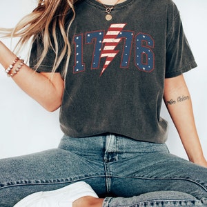 May include: A dark gray t-shirt with a distressed American flag design featuring the number 1776 and a lightning bolt. The shirt is worn by a person wearing blue jeans and white sneakers.