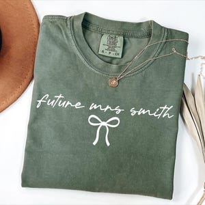May include: A green t-shirt with the text "future mrs smith" in white lettering and a white bow underneath.