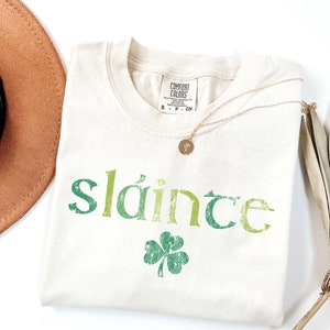 May include: A white t-shirt with a green shamrock and the word "sl&aacute;inte" printed on it in a green, yellow, and white gradient. The shirt is folded and laid flat on a white surface.