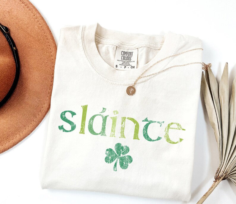 May include: A white t-shirt with a green shamrock and the word "sl&aacute;inte" printed on it in a green, yellow, and white gradient. The shirt is folded and laid flat on a white surface.