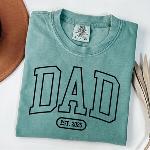 May include: A teal blue t-shirt with the word "DAD" in a black varsity-style font and the text "EST. 2025" in a white circle.