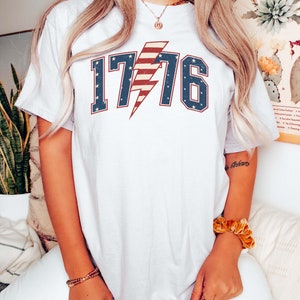 May include: White t-shirt with a red, white, and blue lightning bolt graphic and the number 1776. The graphic is in a vintage style.