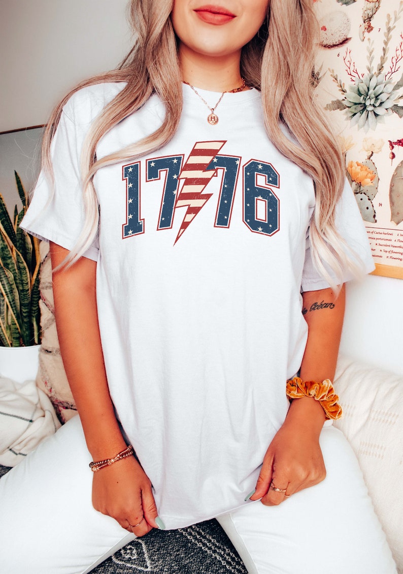 May include: White t-shirt with a red, white, and blue lightning bolt graphic and the number 1776. The graphic is in a vintage style.