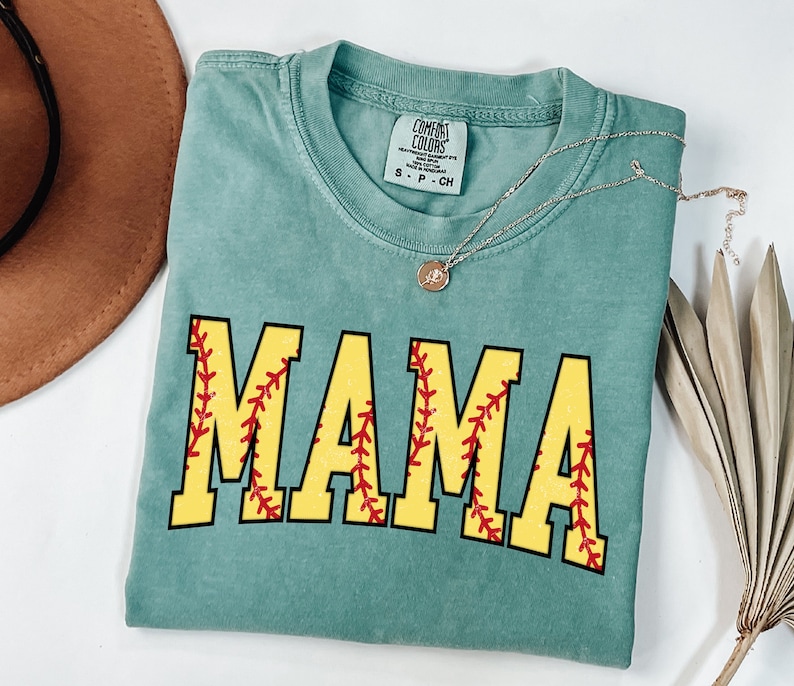 May include: A teal blue t-shirt with the word "MAMA" printed in yellow letters with red stitching, resembling a baseball. The shirt has a distressed look and is made by Comfort Colors.