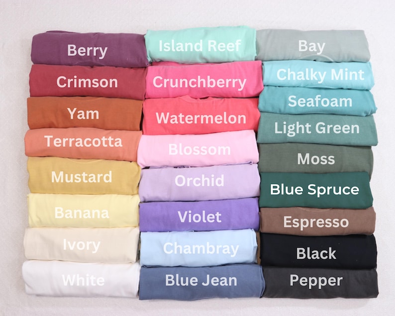 May include: A selection of folded t-shirts in shades like Berry, Crimson, and Yam. Each shirt is labelled with its colour name, including "Island Reef," "Crunchberry," and "Bay." The shirts are neatly stacked, displaying a variety of colours.