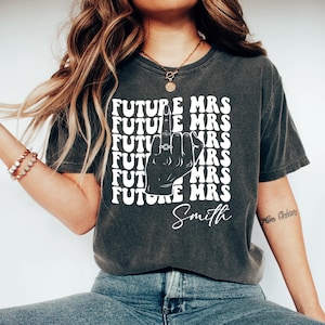 May include: Dark gray t-shirt with the text "FUTURE MRS" repeated in a block, with a hand illustration wearing a ring. The name "Smith" is written below the graphic. The shirt is a casual style.