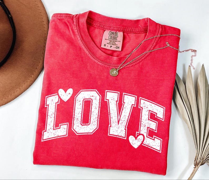 a red t-shirt with the word "LOVE" printed on it, along with two heart symbols. The t-shirt is folded and placed on a white surface, with a brown hat and some leaves visible in the background.