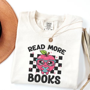 May include: A beige t-shirt with the text "Read More Books" in black. A cartoon apple with glasses and a book is in the center of the text. The apple is pink with a red stem and green leaves.