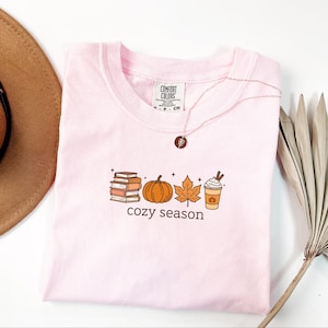 May include: A pale pink t-shirt with a crew neck, displaying a fall-themed graphic. The graphic includes a stack of books, a pumpkin, a leaf, and a coffee cup, with the words "cozy season" below. A gold necklace is draped over the shirt.