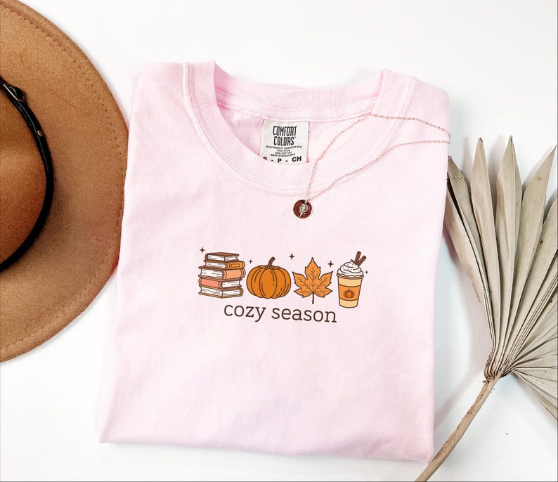 May include: A pale pink t-shirt with a crew neck, displaying a fall-themed graphic. The graphic includes a stack of books, a pumpkin, a leaf, and a coffee cup, with the words "cozy season" below. A gold necklace is draped over the shirt.