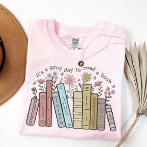 K&ouml;nnte beinhalten: Hellrosa T-Shirt mit dem Aufdruck "it's a good day to read a book". Das Design zeigt bunte B&uuml;cher und Blumen. Eine goldene Halskette und ein brauner Hut sind ebenfalls sichtbar.