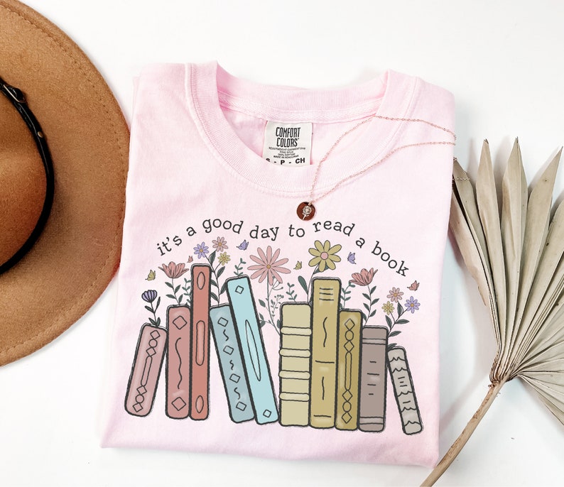K&ouml;nnte beinhalten: Hellrosa T-Shirt mit dem Aufdruck "it's a good day to read a book". Das Design zeigt bunte B&uuml;cher und Blumen. Eine goldene Halskette und ein brauner Hut sind ebenfalls sichtbar.