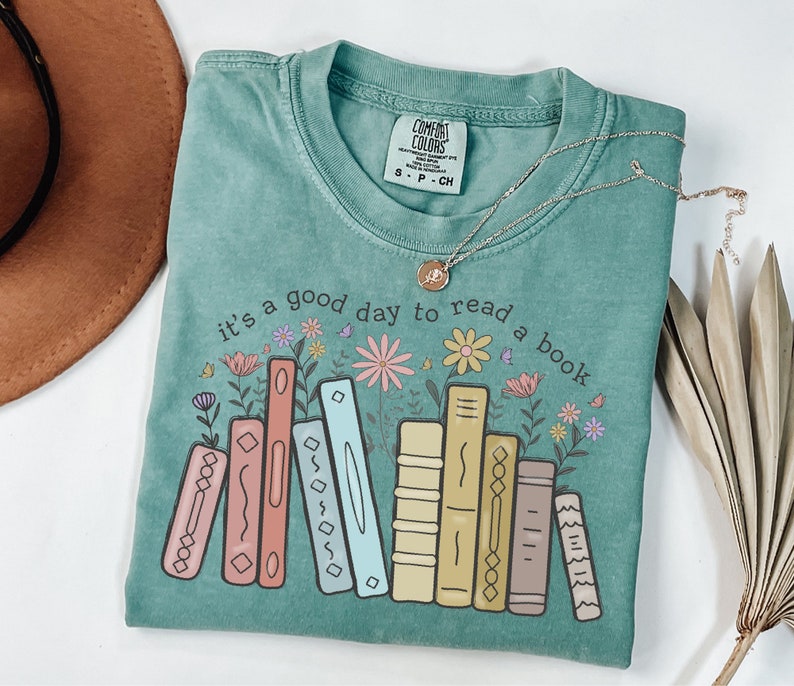 K&ouml;nnte beinhalten: Ein t&uuml;rkisfarbenes Comfort Colors T-Shirt mit dem Schriftzug "it's a good day to read a book" und einer Grafik von bunten B&uuml;chern und Blumen. Eine goldene Halskette mit Anh&auml;nger liegt auf dem Shirt.