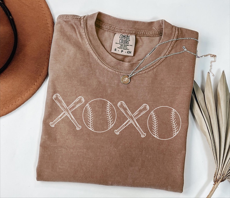 May include: A brown t-shirt with a white graphic of two baseball bats crossed over each other with a baseball on either side of the bats. The graphic says "XOXO".