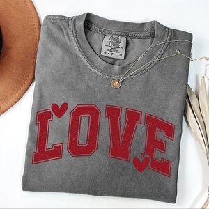 a gray t-shirt with the word "LOVE" printed in red letters, along with two red heart symbols. The t-shirt is placed on a white surface, and there is a brown hat and some dried leaves nearby.