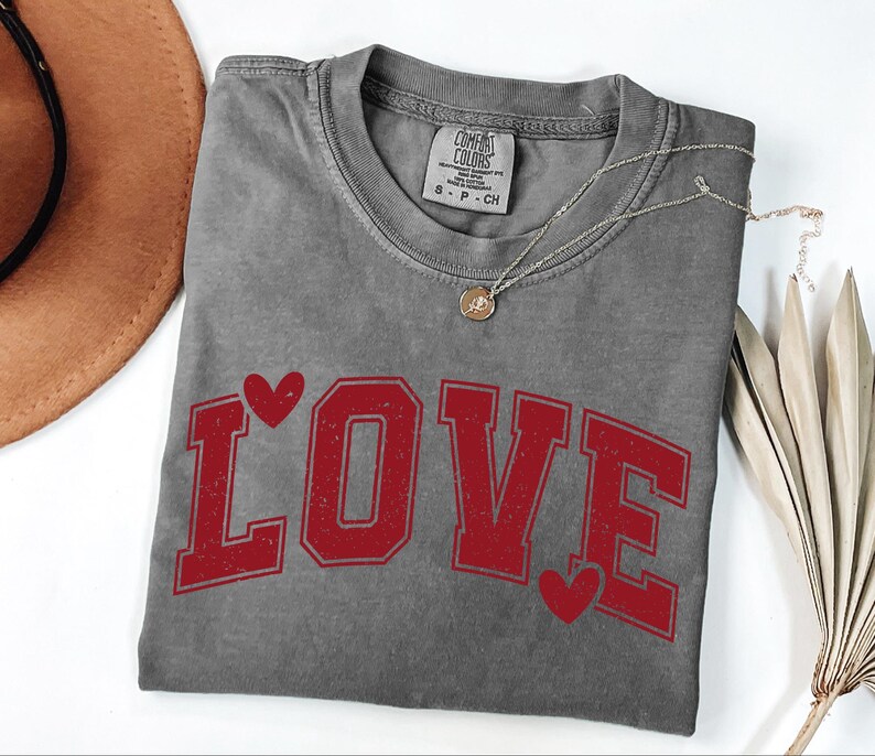 a gray t-shirt with the word "LOVE" printed in red letters, along with two red heart symbols. The t-shirt is placed on a white surface, and there is a brown hat and some dried leaves nearby.
