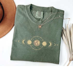 K&ouml;nnte beinhalten: Ein gr&uuml;nes T-Shirt mit einer goldenen Grafik der Erde und Mondphasen. Das Shirt hat den Text "COMFORT COLORS" auf dem Etikett.