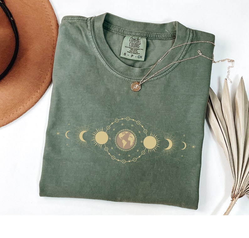 K&ouml;nnte beinhalten: Ein gr&uuml;nes T-Shirt mit einer goldenen Grafik der Erde und Mondphasen. Das Shirt hat den Text "COMFORT COLORS" auf dem Etikett.
