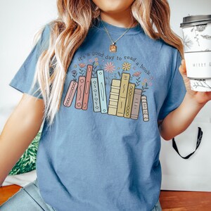K&ouml;nnte beinhalten: Hellblaues T-Shirt mit dem Text "it's a good day to read a book" &uuml;ber einer Grafik von bunten B&uuml;chern und Blumen. Das Design zeigt eine Reihe von B&uuml;chern in Pastellfarben und florale Elemente.