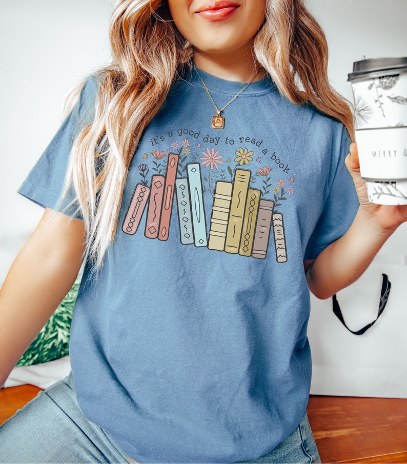 K&ouml;nnte beinhalten: Hellblaues T-Shirt mit dem Text "it's a good day to read a book" &uuml;ber einer Grafik von bunten B&uuml;chern und Blumen. Das Design zeigt eine Reihe von B&uuml;chern in Pastellfarben und florale Elemente.