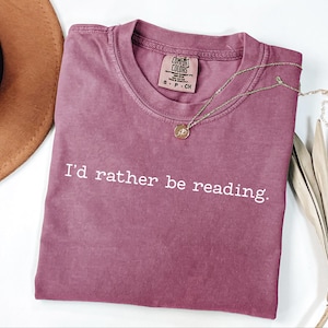 May include: A mauve-colored t-shirt with the text "I'd rather be reading" in white. A gold necklace with a pendant rests on the shirt. A brown hat and dried palm leaves are in the background.