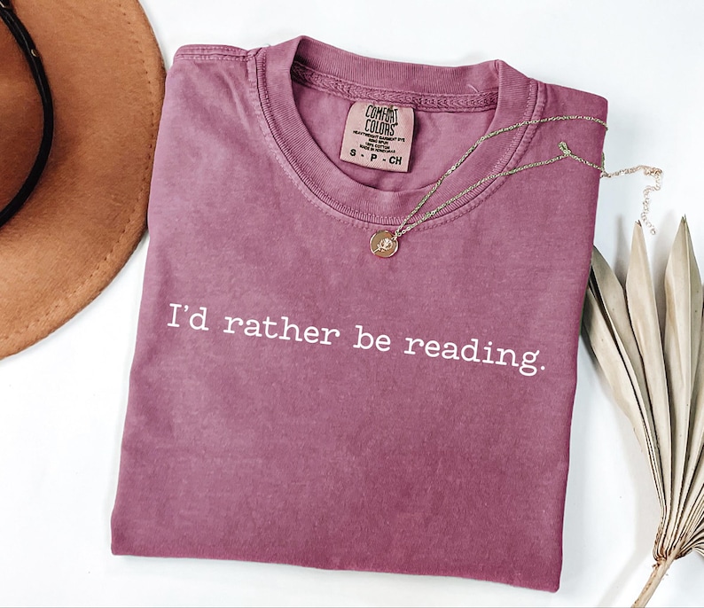May include: A mauve-colored t-shirt with the text "I'd rather be reading" in white. A gold necklace with a pendant rests on the shirt. A brown hat and dried palm leaves are in the background.