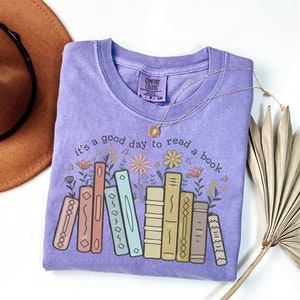 K&ouml;nnte beinhalten: Ein fliederfarbenes Comfort Colors T-Shirt mit dem Schriftzug "it's a good day to read a book" und einer Grafik von bunten B&uuml;chern und Blumen. Eine goldene Halskette und ein brauner Hut sind ebenfalls im Bild.