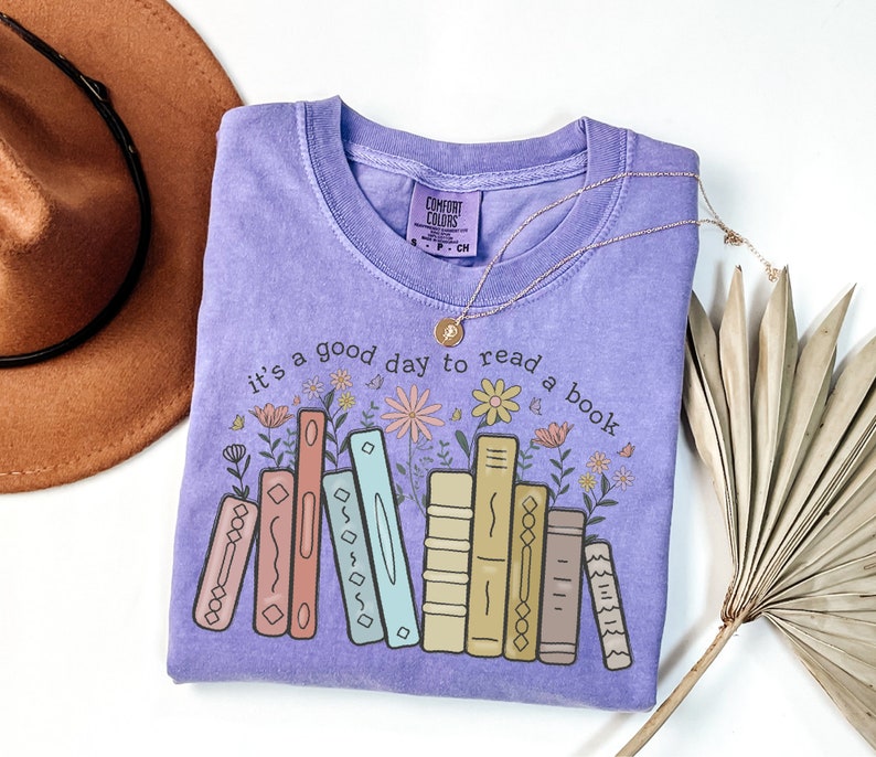 K&ouml;nnte beinhalten: Ein fliederfarbenes Comfort Colors T-Shirt mit dem Schriftzug "it's a good day to read a book" und einer Grafik von bunten B&uuml;chern und Blumen. Eine goldene Halskette und ein brauner Hut sind ebenfalls im Bild.