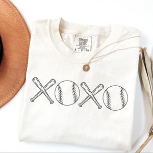 May include: A white t-shirt featuring a black graphic design of two baseball bats crossed over each other with a baseball on either side of the bats. The graphic design reads "XOXO".