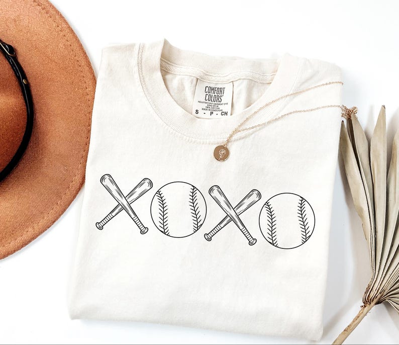 May include: A white t-shirt featuring a black graphic design of two baseball bats crossed over each other with a baseball on either side of the bats. The graphic design reads "XOXO".