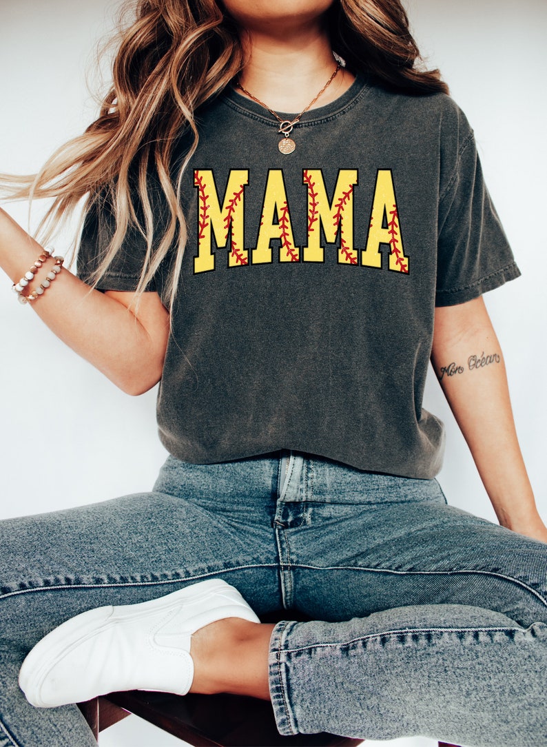 May include: A charcoal grey t-shirt with a yellow softball graphic that says "MAMA" in red stitching.