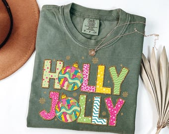 May include: Olive green t-shirt with the words "HOLLY JOLLY" in colorful, patterned letters and Christmas ornament graphics. A silver necklace with a pendant rests on the shirt. The shirt is folded.